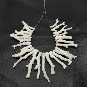 May include: A white coral branch necklace. The necklace is composed of multiple coral branches strung together on a thin, clear string. The branches are a creamy white color and have a natural, organic shape. The necklace is displayed against a black background.