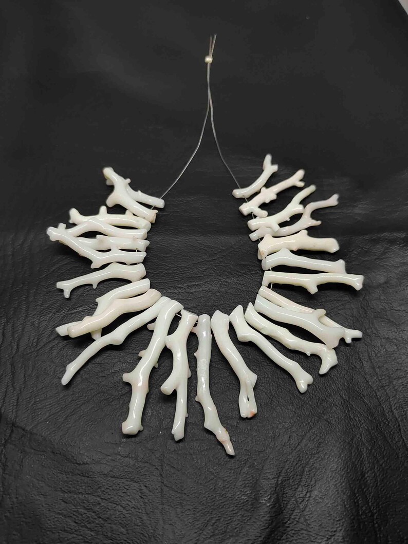 May include: A white coral branch necklace. The necklace is composed of multiple coral branches strung together on a thin, clear string. The branches are a creamy white color and have a natural, organic shape. The necklace is displayed against a black background.