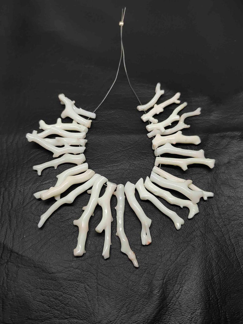 May include: A white coral branch necklace, strung on a thin, clear cord. The coral pieces are irregular in shape, with a natural, organic appearance. The necklace is displayed against a dark background.