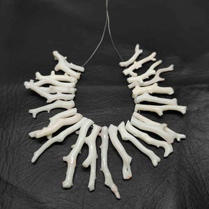 May include: A white coral branch necklace. The necklace is composed of multiple coral branches strung together on a thin, clear cord. The branches are a creamy white color, with some showing subtle pink hues. The background is a dark, textured surface.