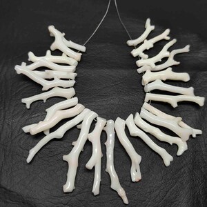 May include: Strung white coral branch beads. The coral pieces have a natural, organic shape, with a slightly textured surface. The beads are strung on a thin, clear cord, and are displayed against a dark background. The coral has a subtle pink hue.