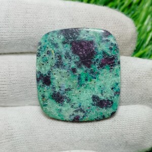 Pode incluir: Uma pedra preciosa retangular polida com um padrão mosqueado de tons verdes e vermelho escuro. A superfície da pedra é lisa e as cores são distribuídas em um design orgânico e natural. A gema é segurada em uma luva branca.