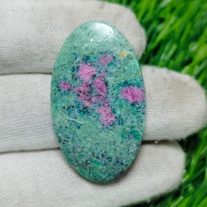 Peut inclure: Pierre précieuse verte de forme ovale avec des inclusions roses et bleues. La pierre précieuse est polie et a une surface lisse.