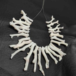 May include: A necklace composed of white coral branches, strung together on a thin, clear cord. The coral pieces have a natural, organic shape, with a slightly textured surface. The background is a dark, solid color.