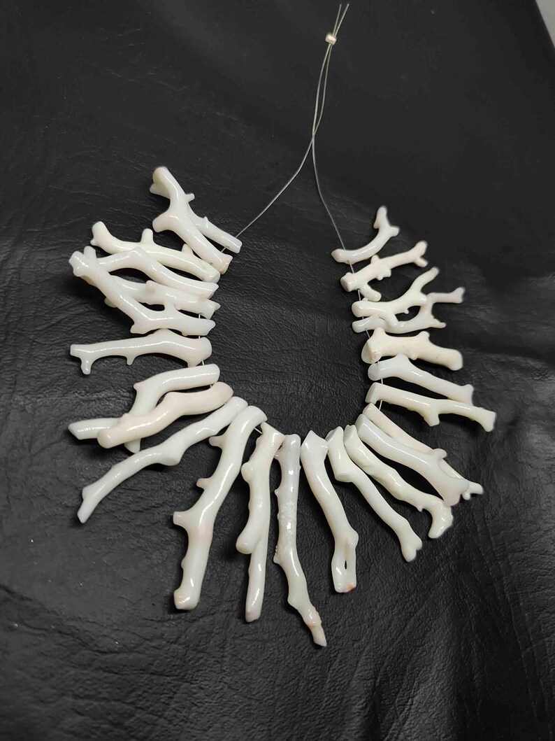 May include: A necklace composed of white coral branches, strung together on a thin, clear cord. The coral pieces have a natural, organic shape, with a slightly textured surface. The background is a dark, solid color.