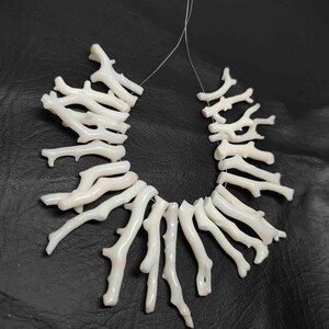 May include: A necklace composed of white coral branches, strung together on a thin, clear cord. The coral pieces have a natural, organic shape, with a slightly textured surface. The background is a dark, solid color.