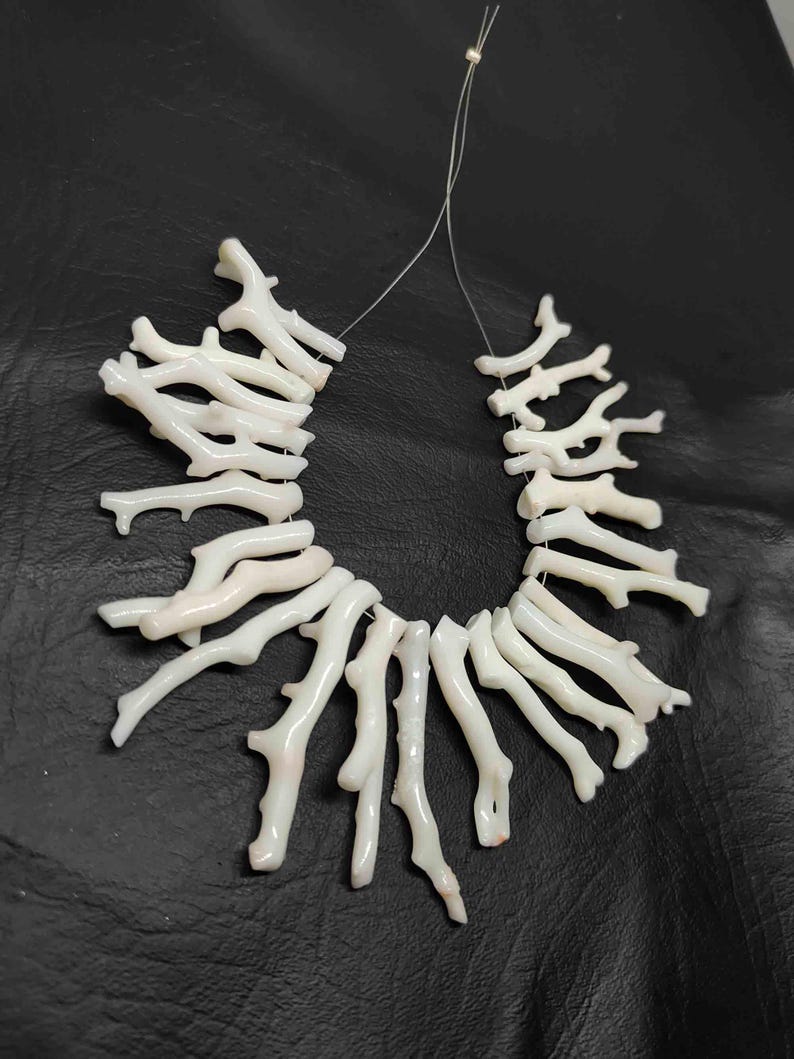 May include: A necklace composed of white coral branches, strung together on a thin, clear cord. The coral pieces have a natural, organic shape, with a slightly textured surface. The background is a dark, solid color.
