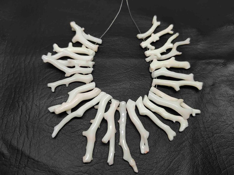 May include: Strands of white coral beads, resembling small branches, are displayed against a dark background. The coral pieces are strung together, showcasing their natural, organic shapes. The beads are a creamy white color.