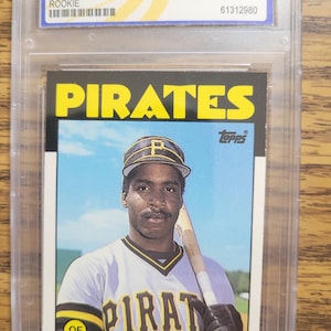 May include: A 1986 Topps baseball card featuring Barry Bonds in a Pittsburgh Pirates uniform. The card is graded WCG 10 GEM-MT and encased in a protective holder. The card's design includes the Pirates logo and Bonds' name.