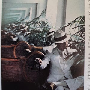May include: A group of people wearing white suits and fedora hats, seated on a wicker bench. The scene is set outdoors with tropical plants in the background. The image has a vintage, possibly retro, aesthetic.
