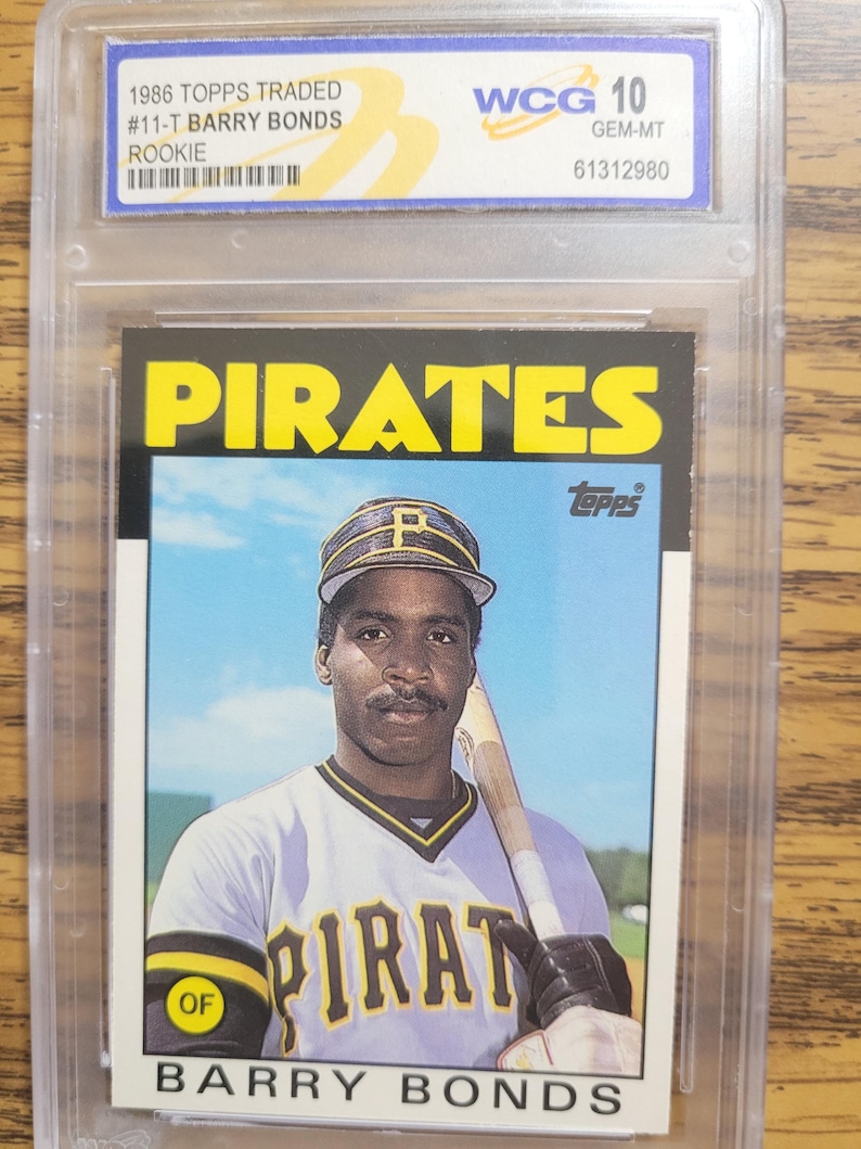 May include: A 1986 Topps baseball card featuring Barry Bonds in a Pittsburgh Pirates uniform. The card is graded WCG 10 GEM-MT and encased in a protective holder. The card's design includes the Pirates logo and Bonds' name.