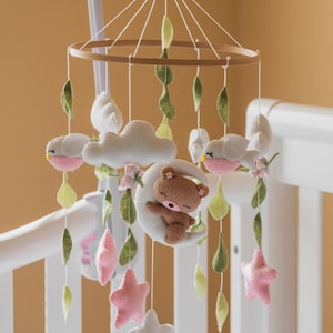May include: A baby mobile with a brown bear sleeping on a crescent moon, two pink and white stars, two white birds with pink accents, and green leaves hanging from a wooden ring.