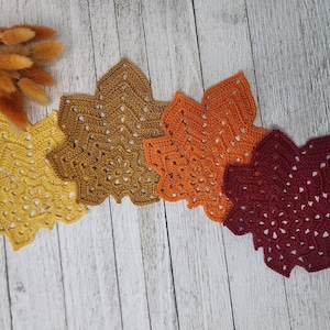 May include: Four crocheted leaf-shaped coasters in a gradient of autumn colours, including mustard yellow, golden brown, burnt orange, and deep burgundy. Each coaster features an intricate, openwork lace pattern with a central floral motif.