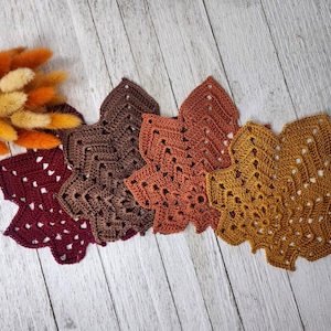 May include: Four crocheted leaf-shaped doilies in shades of burgundy, dark brown, burnt orange, and mustard yellow, arranged in a row next to a small cluster of dried orange and cream-coloured bunny tail grass stems.
