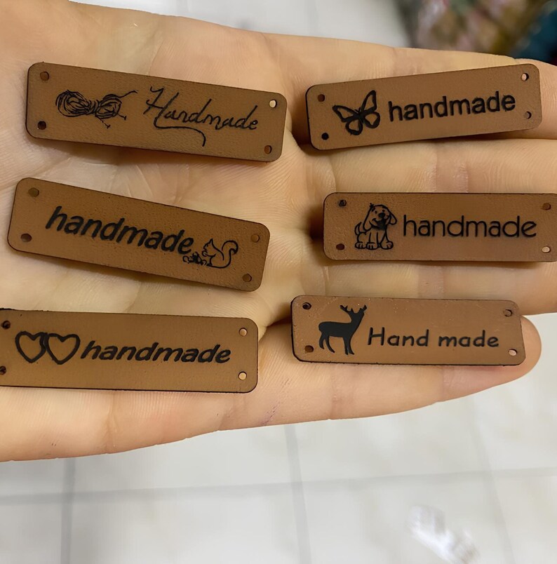 May include: Six brown leather labels with different designs and the word "handmade" written on them. The designs include a butterfly, a squirrel, a dog, a deer, a heart, and a ball of yarn.
