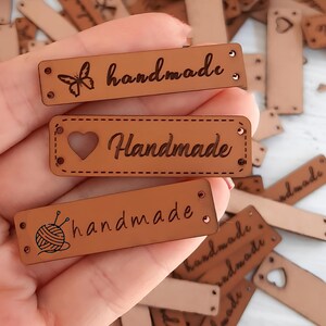 May include: Three brown leather labels with the word "handmade" written in cursive. The labels have different designs: a butterfly, a heart, and a ball of yarn with knitting needles.
