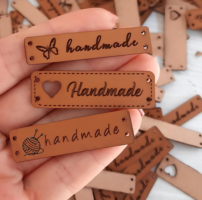 May include: Three brown leather labels with the word "handmade" written in cursive. The labels have different designs: a butterfly, a heart, and a ball of yarn with knitting needles.