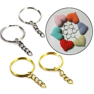 May include: Four keychains with silver and gold rings and chains. The keychains are shown next to a circle containing six crocheted heart-shaped keychains in pink, orange, blue, turquoise, red, and beige.
