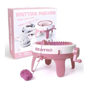 Può includere: Una macchina da maglia rosa e bianca con una manovella. La macchina è su una superficie bianca con un gomitolo di filato rosa accanto. La scatola della macchina è sullo sfondo e dice "Knitting Machine Magic Loop Loom More Than a Toy".