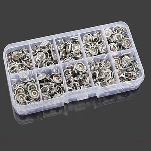 May include: A clear plastic storage box filled with silver metal snap fasteners. The box has nine compartments, each filled with different sizes of snaps.