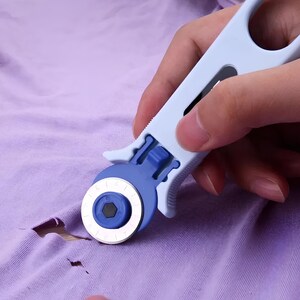 May include: A blue and white rotary fabric cutter with a safety guard. The cutter is being used to cut a hole in a purple fabric.