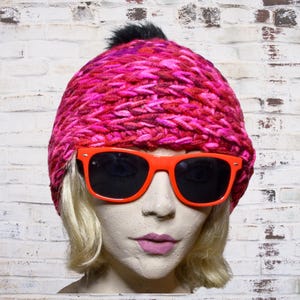 May include: A mannequin head wearing a vibrant pink knit hat with a black pom-pom and bright orange sunglasses with dark lenses. The hat is a chunky knit with shades of pink and red.