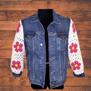 May include: A blue denim jacket with white crochet sleeves. The crochet sleeves feature pink flowers with yellow centres. The jacket has a classic collar, button closures, and pockets. The jacket is displayed on a black mannequin.