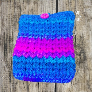 May include: Handmade crochet sleeve in shades of blue and magenta, secured with a pink button. The sleeve features a textured stitch pattern and is designed to hold a small item. The item is placed on a weathered wooden surface.