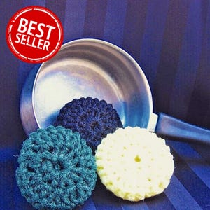 May include: Three crocheted dish scrubbers in teal, navy blue, and yellow, beside a stainless steel pan. The scrubbers are round with a textured surface. A red circle with the words "BEST SELLER" is in the upper left corner.