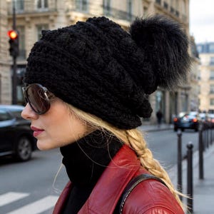 May include: A black crocheted beanie featuring a textured bobble stitch pattern and a large, fluffy black faux fur pom-pom on top. The hat is constructed from thick yarn and provides full coverage for the head with a slightly slouchy fit.