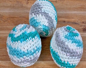May include: Three crocheted balls in a teal, white, and gray color pattern. The balls are made with a textured yarn and have a slightly bumpy surface.