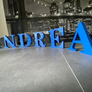 Könnte beinhalten: Blaue Holzbuchstaben, die den Namen "ANDREA" auf einer grauen Oberfläche buchstabieren. Die Buchstaben stehen frei und haben eine leicht strukturierte Oberfläche.