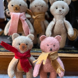 Op de afbeelding: Vijf handgemaakte vilten teddyberen in verschillende tinten grijs en roze. Elke beer heeft een kleine sjaal in kleuren als roze, rood en goud. De beren staan voor een vintage naaimachine.