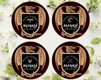 May include: Four round coasters with a dark brown background and gold accents. Each coaster features a dragon with wings spread out and a shield with the text "BASGIATH WAR COLLEGE" and a different quadrant name: Riders, Scribes, Healers, and Infantry.