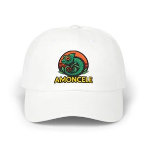 Peut inclure: Une casquette de baseball blanche ornée d'un motif circulaire représentant un caméléon vert aux yeux orange sur un fond de coucher de soleil orange. Sous le motif, le mot AMONCELE est imprimé en lettres capitales jaunes à contour noir.