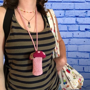 May include: A pink and burgundy crochet mushroom pendant necklace. The mushroom has a pink stem and a burgundy cap with white polka dots. The necklace is worn over a striped tank top.