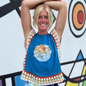 May include: A blue t-shirt with the text "Lefty Lucy" and a sun and wave design, paired with a colorful crochet top. The crochet features a granny square design with orange, white, and blue accents.