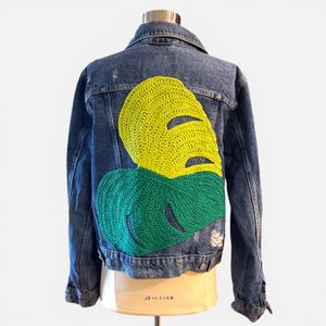 May include: A blue denim jacket with a large, two-tone crochet leaf design on the back. The leaf is yellow and green, with a distressed look. The jacket has a classic collar and button closure.
