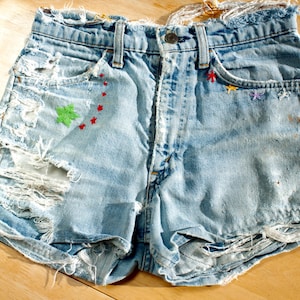 May include: Light blue denim shorts with distressed detailing, featuring multiple rips and embroidered accents. The shorts have a button closure and pockets, with colourful embroidered stars and a green leaf design.