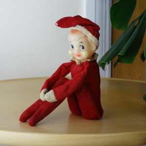 Könnte beinhalten: Eine Vintage-Elf-Puppe mit rotem Filz-Outfit und -Hut, cremefarbenen Verzierungen und einem bemalten Gesicht. Die Puppe sitzt mit verschränkten Beinen, Armen auf den Knien und einem kleinen Rüschenkragen.
