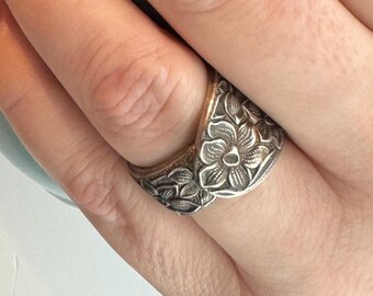 May include: Silver-toned ring with a floral design. The ring features detailed carvings of flowers and leaves, creating a textured surface. The ring is worn on a finger, showcasing the intricate craftsmanship and design.