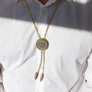 May include: A gold-coloured bolo tie with a silver coin pendant featuring a profile. The coin is encircled by a twisted rope design, and the tie has a braided cord with decorative metal tips. The tie is worn over a white shirt.