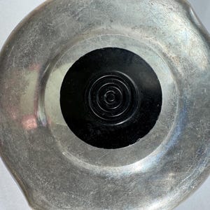 Könnte beinhalten: Ein silberfarbenes, kreisförmiges Objekt mit einem schwarzen, spiralförmigen Zentrum. Die Metalloberfläche weist Gebrauchsspuren auf, die auf Alter oder Nutzung hindeuten. Das Design ist einfach und funktional, möglicherweise ein Deckel oder Behälter.