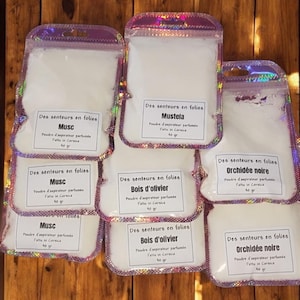 以下が含まれることがあります： ピンクと虹色の縁取りが付いた小さな白い袋が8つ。各袋には白い粉末が入っており、「Des senteurs en folies」というテキストと、フランス語で異なる香りの名前が書かれたラベルが付いています。香りは、ムスク、ムステラ、オリーブの木、黒い蘭です。