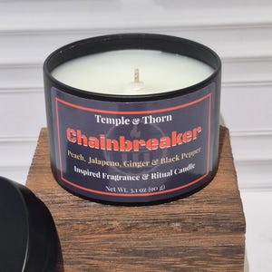 May include: A black metal candle tin with a white wax fill and a single wick. The label reads Temple & Thorn Chainbreaker, Peach, Jalapeno, Ginger & Black Pepper Inspired Fragrance & Ritual Candle, Net Wt. 3.1 oz (90 g). A matching black lid sits nearby.