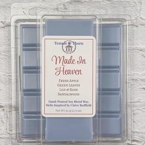 May include: A light blue wax melt bar in a clear plastic clamshell container. The label reads Temple & Thorn, Made In Heaven, Fresh Apple, Green Leaves, Lily & Rose, Sandalwood, Hand-Poured Soy Blend Wax Melts Inspired by Claire Redfield, Net Wt 1.75 oz.