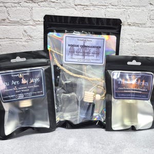 May include: Three black pouches containing car diffuser sets. The left pouch is labeled You Are My Hope, the center pouch is labeled Rogue Commander, and the right pouch is labeled The Veteran. Each pouch contains a glass bottle with a wooden cap.