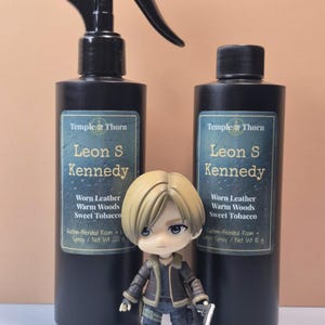 May include: Two black room spray bottles with labels reading "Leon S Kennedy" and a small figurine of a character holding a gun. The labels list "Worn Leather, Warm Woods, Sweet Tobacco".