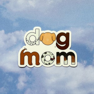 Peut inclure: Autocollant blanc avec le texte "dog mom" en lettres brunes et noires. Les lettres sont décorées de silhouettes de chiens.