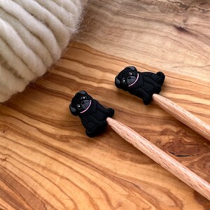 May include: Two wooden knitting needles with black pug dog-shaped stitch markers. The stitch markers have pink collars.